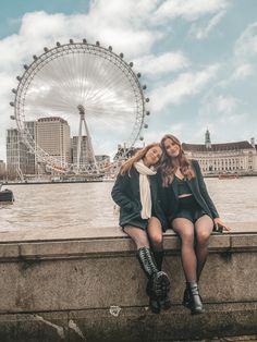 two women are sitting on a wall near the water and a ferris wheel in the background