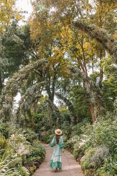 a woman wearing a hat walks down a path in the middle of trees and bushes