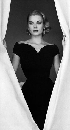 a black and white photo of a woman in a dress looking out from behind a curtain