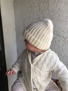 a baby sitting on the ground wearing a white sweater and hat, with one foot in the air