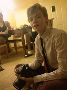 a young man sitting on the floor with a camera in his hand and two other people behind him