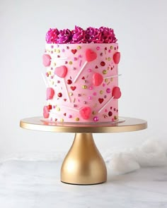 a pink cake decorated with hearts and sprinkles on a gold plated stand