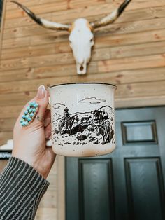 a person holding up a coffee mug with a cow skull on the wall behind it