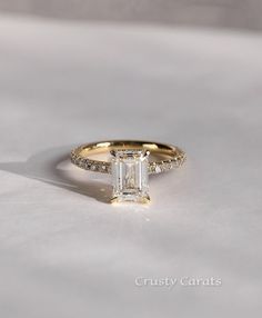 a gold ring with an emerald cut diamond surrounded by pave diamonds on a white surface