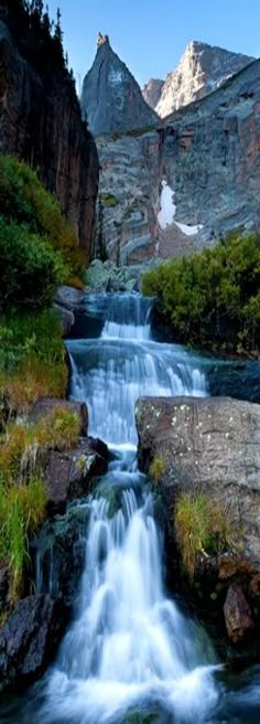 a small waterfall running through a lush green forest filled with rocks and grass next to tall mountains