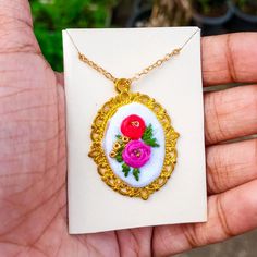 a hand holding a necklace with flowers on it in a gold frame and white background