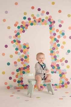 a baby sitting on a stool in front of a wall with multicolored confetti