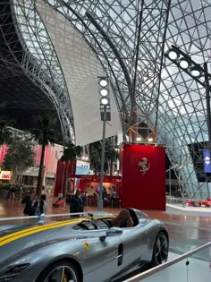 a silver sports car parked in front of a large glass building with people walking around it