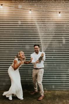 a man and woman standing next to each other holding champagne bottles in front of a metal wall