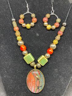 Unique Fluid art pendant and earrings, featuring greens and red in antique brass setting . This pendant is made from acrylic paint that dripped from my pour paintings. The paint dries and makes a skin which is positioned under a glass cabochon and then placed into the nickel free stainless steel bezel. The statement piece is in shades of red and green and the beads complement the colors, all in an antique brass setting. Earrings match. Color is my passion and each is unique in its own way. Beads are glass and plastic and the cord is wax coated. The length is adjustable with a sliding knot. Acrylic Pour Jewelry, Artsy Hand Painted Copper Jewelry, Multicolor Hand Painted Metal Jewelry, Handmade Artsy Bronze Jewelry, Hand Painted Multicolor Metal Jewelry, Artsy Handmade Bronze Jewelry, Bronze Jewelry With Artistic Design, Unique Multicolor Brass Jewelry, Artistic Bronze Jewelry With Artistic Design