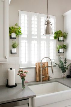 a kitchen sink sitting under a window next to a wooden cutting board and potted plants