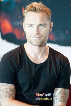 a man with tattoos sitting in front of a wall and looking at the camera while wearing a black t - shirt