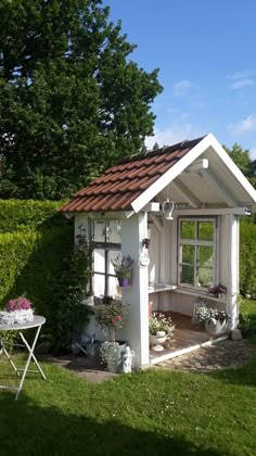 a small white shed with potted plants on the side and a table in front