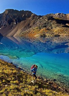 a person with a backpack walking up a hill next to a body of water and mountains