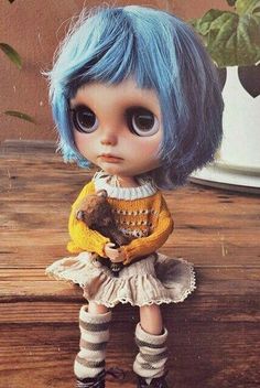 a doll with blue hair is sitting on a wooden table next to a potted plant