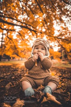 a baby sitting on the ground in front of a tree with autumn leaves around it