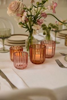 the table is set with pink glasses and flowers in vases, plates and silverware