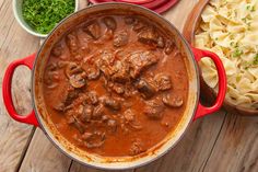 a red pot filled with meat and pasta on top of a wooden table next to other dishes