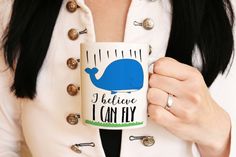 a woman holding a coffee mug with a blue whale on it's side that says i believe i can fly