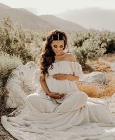 a pregnant woman sitting in the desert with her hands on her stomach and wearing a white dress