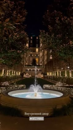 an illuminated fountain in front of a building at night with trees and bushes around it