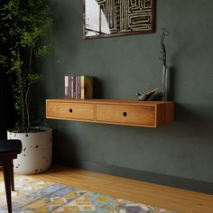 A modern room showcases a Krovel Floating Desk in Cherry with two drawers. On top, books and a vase sit elegantly. The dark green wall features abstract art, complemented by a nearby large potted plant. A colorful rug on the wooden floor highlights handmade details throughout. Floating Furniture, Floating Desks, White Oak Furniture, Maple Furniture, Cherry Furniture, Dresser With Tv, Media Console Table, Floating Cabinets, Floating Desk