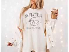 a woman holding a drink in her right hand and wearing a newweds club t - shirt