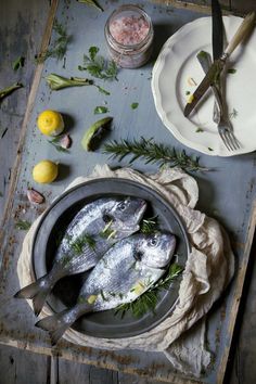 two fish are sitting in a pan on a table next to lemons and herbs