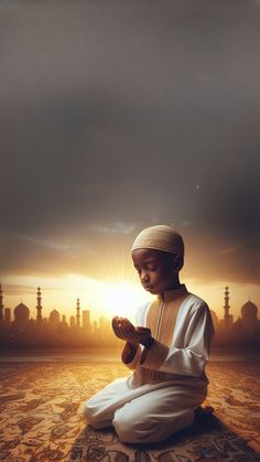 a young boy sitting on the ground in front of a sunset with his hands together