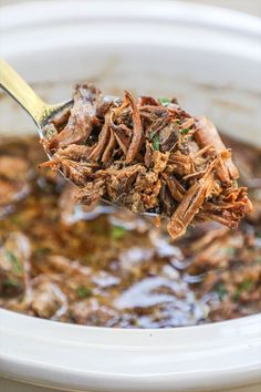 a spoon full of pulled pork and brown gravy in a white crock pot