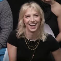 a woman with blonde hair is smiling and wearing gold chains on her neck, while two other women are in the background