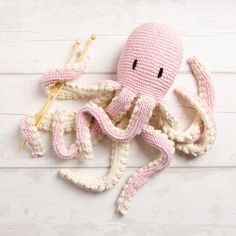 an octopus crocheted in pink and white sits next to knitting needles on a wooden surface