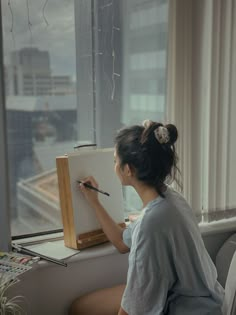 a woman sitting in front of a window writing on a white board with a brush