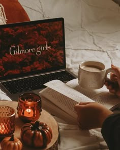 a person sitting on a bed reading a book and drinking coffee while using a laptop