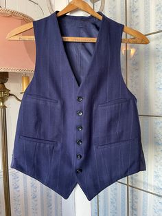 This tailored vest is made from a wool blend in dark royal/navy blue, with widely spaced light blue and beige pinstripes. It has the original buttons in the front and four front pockets, and is fully lined. There are no labels. The measurements, taken with the vest lying flat. are: shoulder to shoulder, 11 inches; armpit to armpit, 19 inches; length, 24 inches in front and 20 inches in back; bottom edge, 17 inches. In very good condition. Navy Sleeveless Vest With Pockets, Classic Blue Three-piece Suit With Pockets, Classic Blue Three-piece Suit, Wool Vest With Pockets For Tailoring, Fitted Navy Sleeveless Vest, Fitted Pinstripe Outerwear With Buttons, Elegant Tailored Pinstripe Vest, Navy Fitted Vest For Workwear, Elegant Wool Vest With Pockets