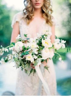 a woman in a wedding dress holding a bridal bouquet with white flowers and greenery