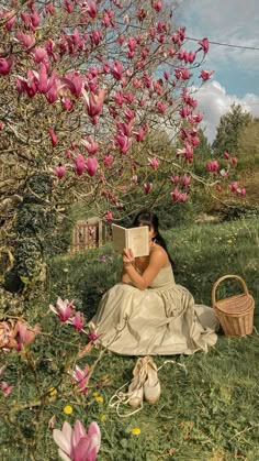 a woman sitting in the grass reading a book under a tree filled with pink flowers