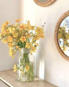 yellow flowers are in a clear vase on a wooden table next to two round mirrors