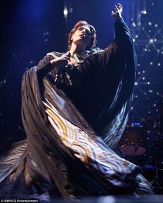 a woman in a black dress on stage with her arms out and one hand up