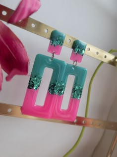 two pink and blue earrings hanging from a metal hook with flowers in the back ground