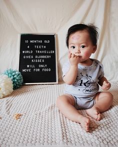 a baby sitting on a bed eating food next to a sign that says, 10 months old