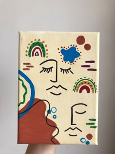 a person holding up a small painting with faces on it's face and rainbows in the background