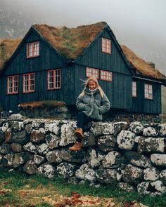 a woman sitting on a stone wall in front of a black house with grass roofs