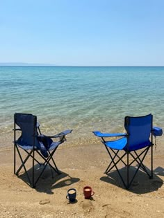 two blue chairs sitting on top of a sandy beach next to the ocean and a bucket