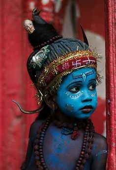 a young boy with blue face paint and headdress, standing in front of a red curtain