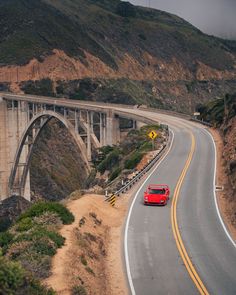 a red car is driving down the road near a bridge and mountain side with hills in the background