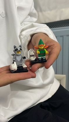 a person holding two lego minis in their hands, one is wearing a white shirt and the other has a green hat