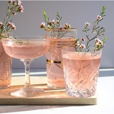 three glasses filled with pink liquid sitting on top of a wooden tray next to flowers