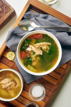 a bowl of chicken and vegetable soup on a wooden tray with two spoons next to it