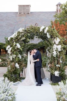 a bride and groom kissing in front of an outdoor wedding ceremony arch with white flowers